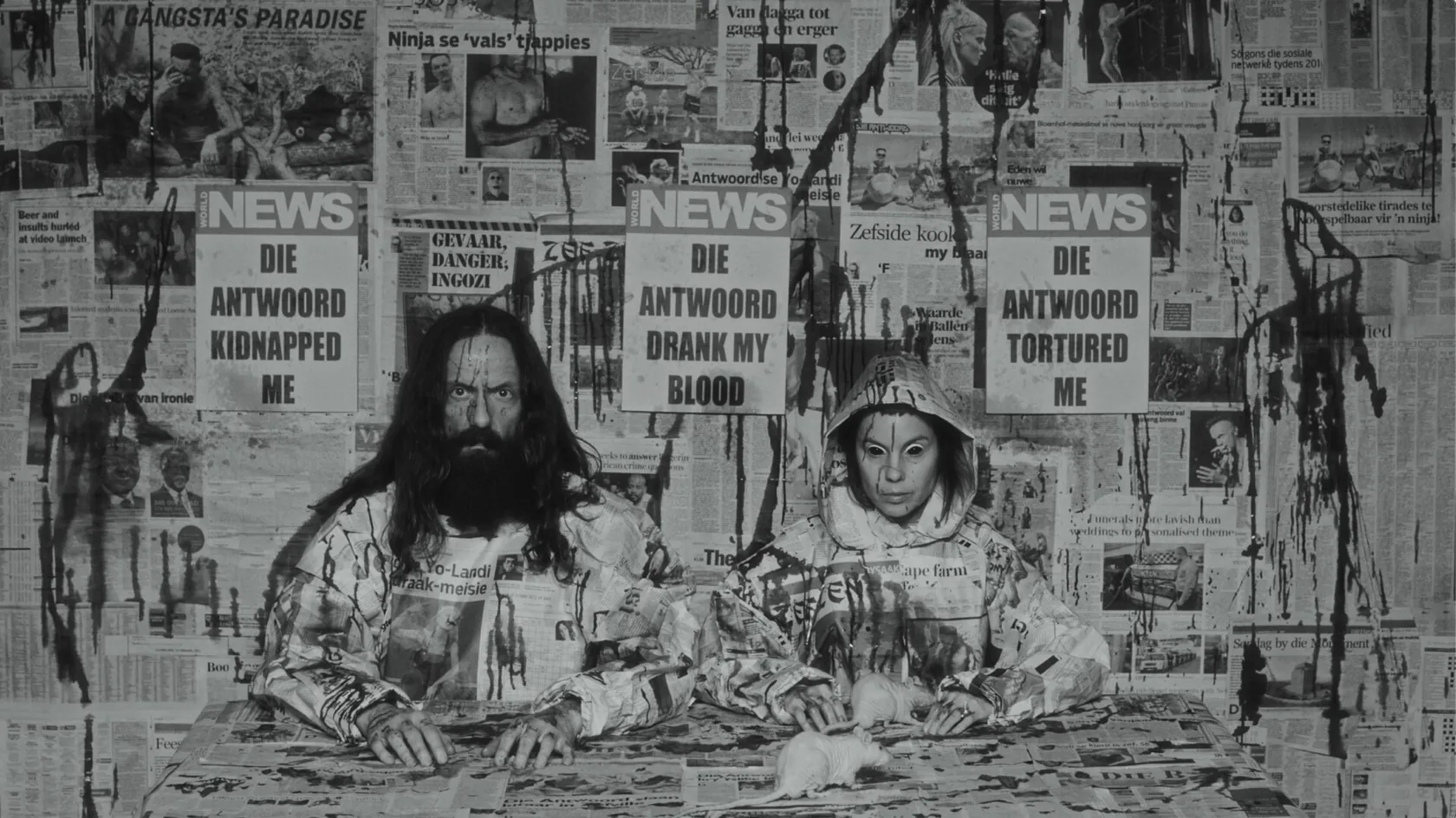 Two figures seated before a wall of stark newspaper headlines.